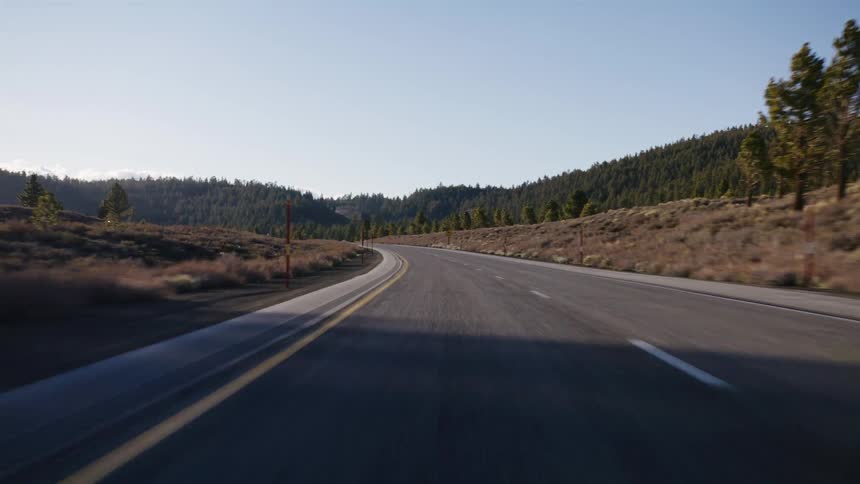 A first-person view driving along a winding, empty highway through a scenic mountain forest on a sunny day