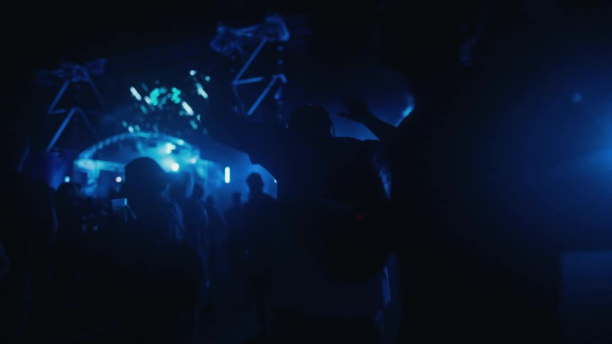 Silhouetted person with arms outstretched dances in the crowd at a nighttime music festival with vibrant blue stage lights