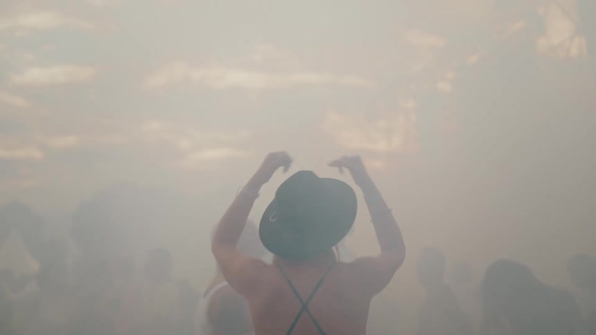 A slow-motion upward tilt follows a woman in a hat as she dances with a crowd in a thick cloud of smoke at a music festival