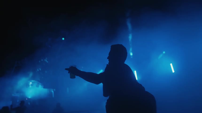 A man's silhouette is seen dancing and talking on the phone at a music festival with blue lights and smoke