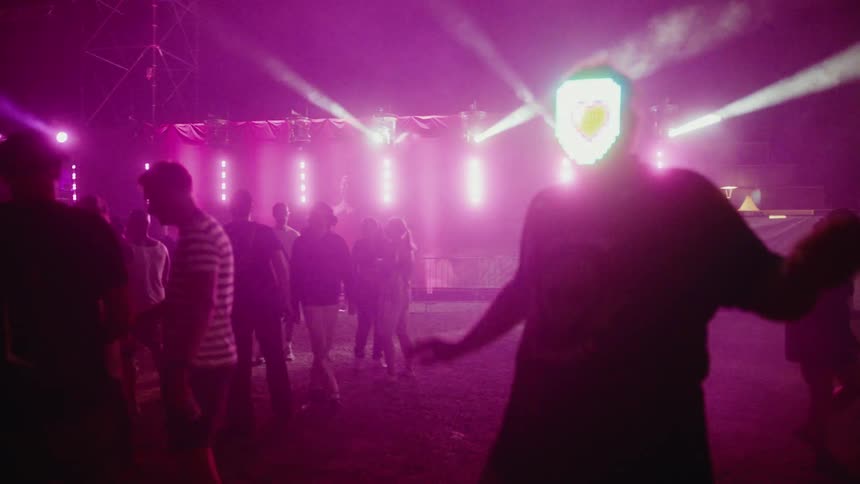 A man in a futuristic LED mask dances and interacts with the camera at a vibrant nighttime music festival with pink stage lights