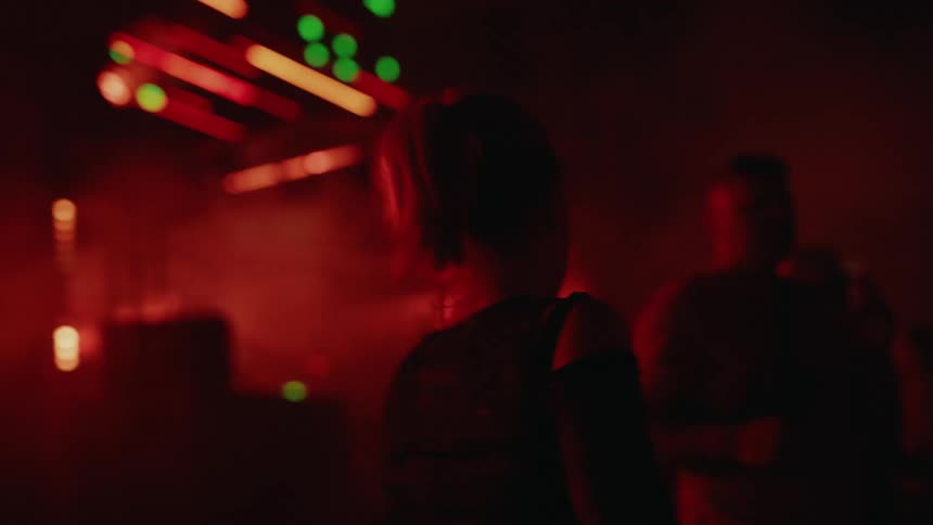 A woman with a ponytail dances in silhouette in a dark, red-lit nightclub filled with smoke and a crowd of people