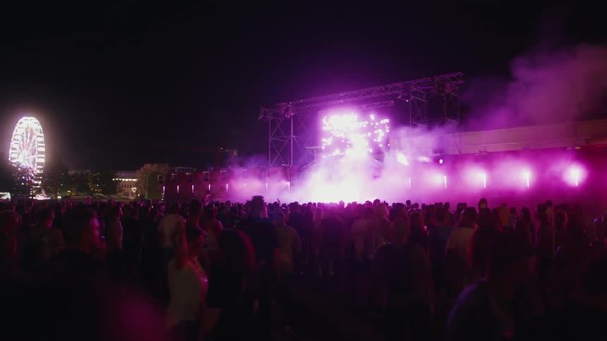 An enthusiastic crowd dances at a vibrant outdoor music festival at night, with the stage erupting in purple lights and smoke effects