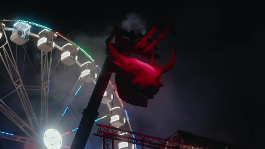 A large bull's head sculpture is illuminated in red light in front of a colorful spinning Ferris wheel at a nighttime amusement park