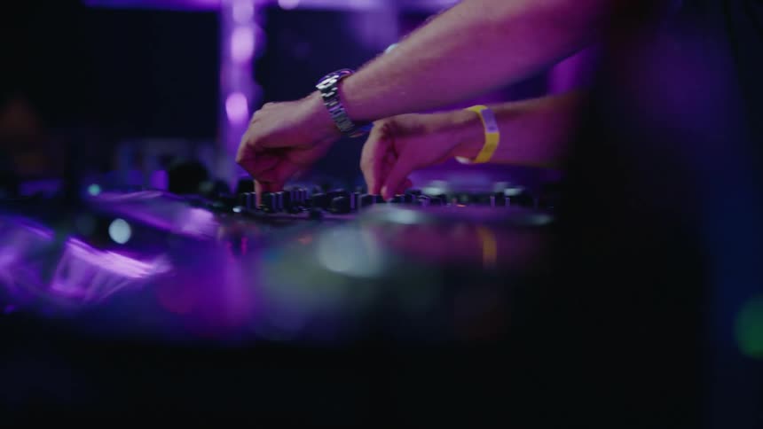 A DJ's hands skillfully adjust the knobs and faders on a mixer during a vibrant, purple-lit set in a nightclub