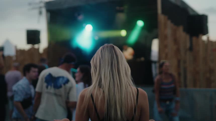 A young blonde woman with her back to the camera dances with her hands in the air at an outdoor music festival