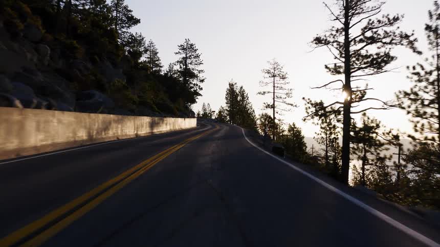 A first-person view of a scenic drive along a winding mountain road as the sun sets over a glistening lake
