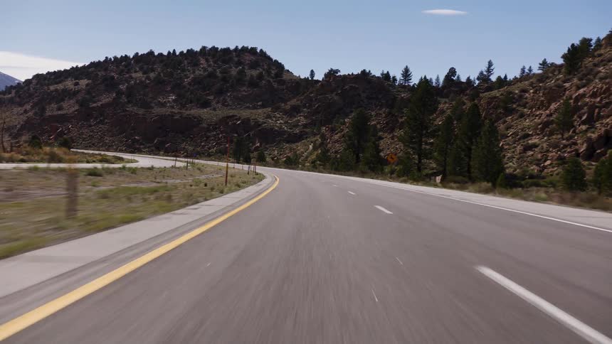 A point-of-view shot driving along a winding highway through a rocky, mountainous landscape under a clear blue sky