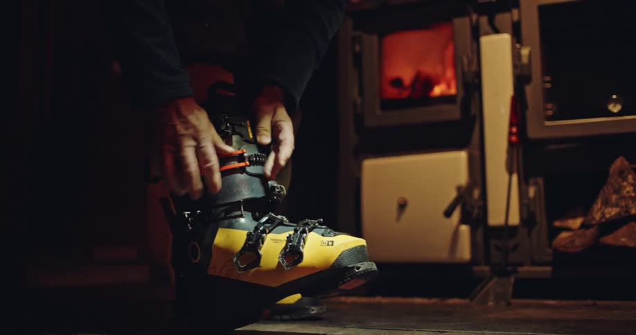 Close up of a skier fastening bright yellow ski boots by the warm glow of a crackling fire in a rustic cabin