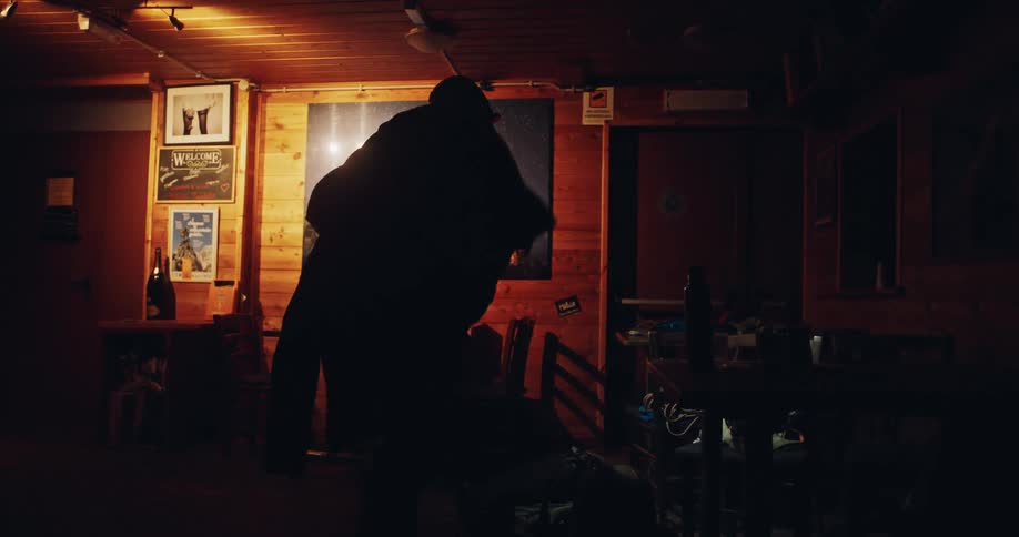A silhouetted man puts on a warm winter jacket inside a cozy mountain lodge, preparing for a cold expedition