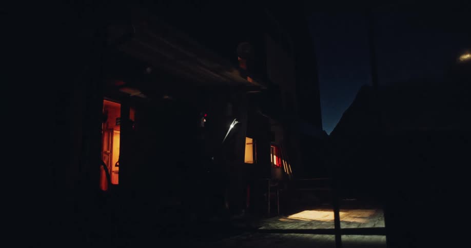 A lone skier with a bright headlamp exits a rustic mountain lodge in the pre-dawn darkness, preparing for an early morning on the slopes