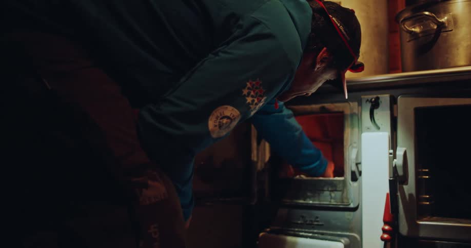 A person wearing a headlamp carefully adds wood and kindling to a vintage stove, stoking a warm fire in a rustic cabin kitchen