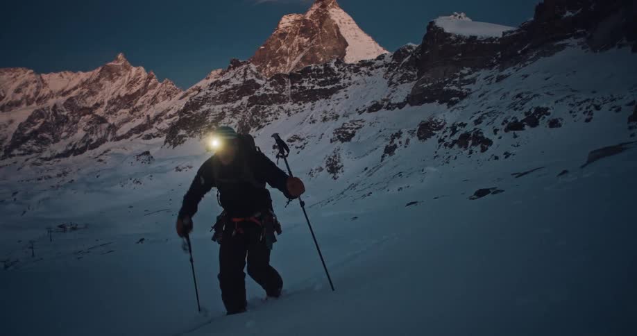 A lone mountaineer with a headlamp on ski tours up a snowy mountain at dawn with the iconic Matterhorn in the background