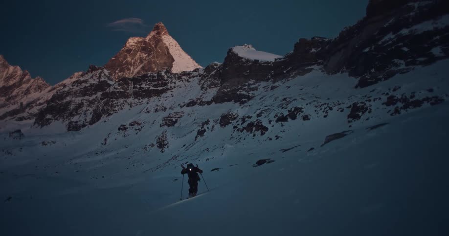 A lone ski mountaineer ascends a snowy slope with a headlamp during the blue hour, with the iconic Matterhorn peak in the background