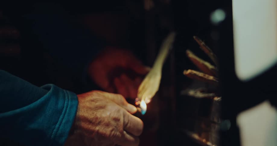 A slow-motion close-up shows an older man with a headlamp using a lighter to ignite kindling in a wood-burning stove