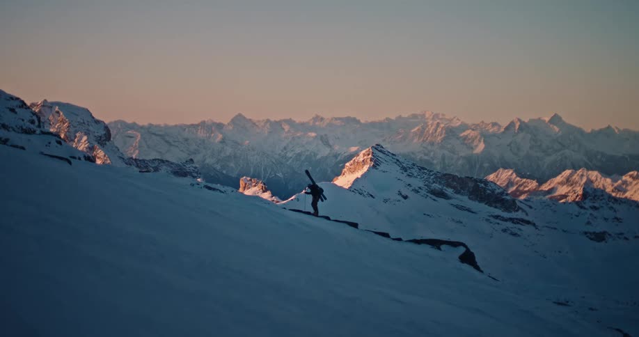 A lone skier hikes up a snowy mountain ridge with their skis on their back as the sun rises over the vast mountain range