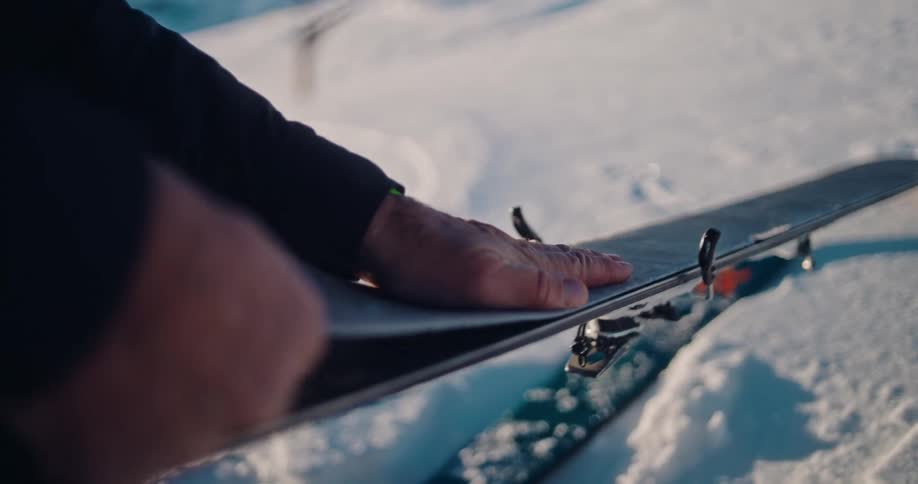 A skier carefully removes the climbing skin from their ski in the bright snow, captured in a detailed slow-motion shot