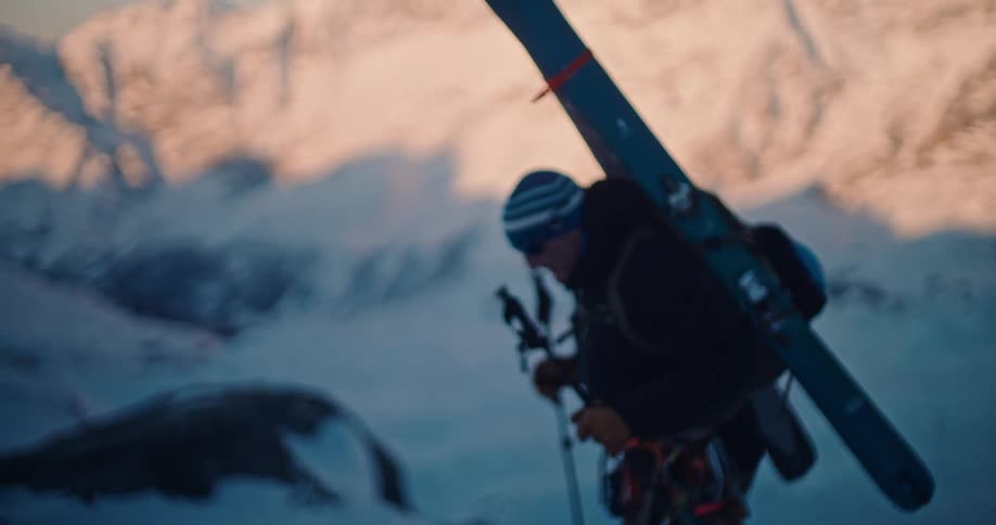 A determined backcountry skier hikes up a steep, snowy mountain slope with his skis on his back during a beautiful sunrise