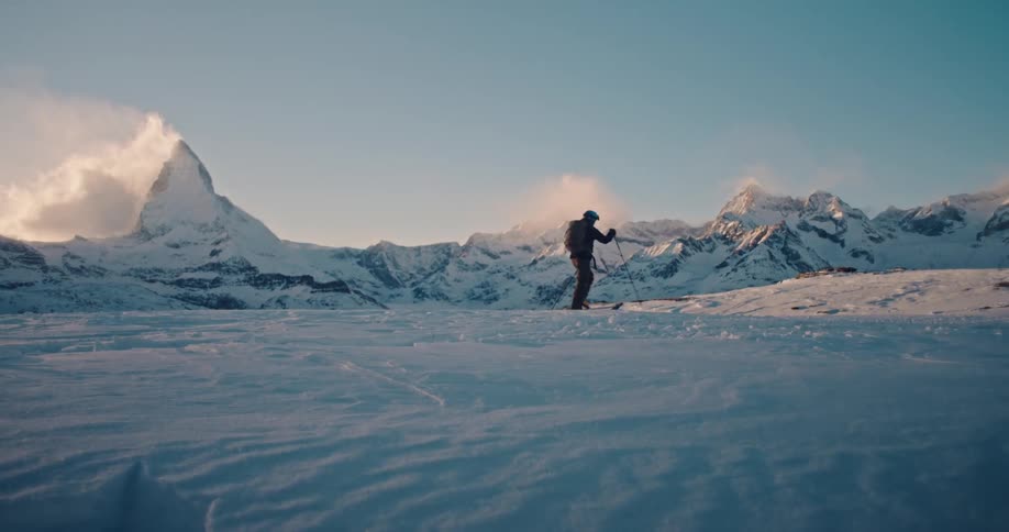 A lone adventurer hikes in slow motion across a vast snowy landscape with the iconic Matterhorn mountain in the background at sunrise