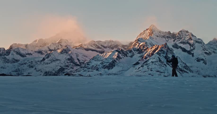 A lone ski tourer hikes across a snowy ridge against a backdrop of majestic, sunlit mountain peaks at sunrise