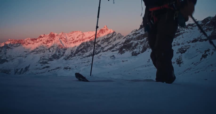 A solitary ski mountaineer skins up a snowy mountain at dawn with the iconic Matterhorn illuminated by a stunning alpenglow in the background