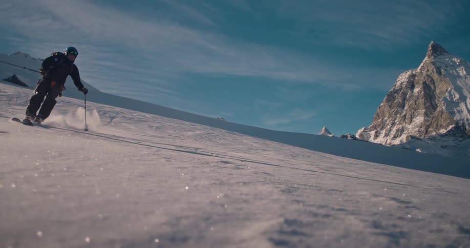 A skier carves through fresh powder in slow motion, spraying snow towards the camera with the magnificent Matterhorn in the background