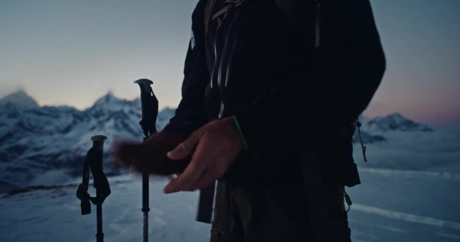 A skier carefully puts on his gloves in slow motion at dawn, preparing for an adventure in a majestic, snow-covered mountain range