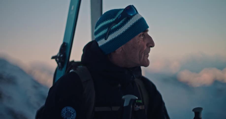 A senior skier hikes up a snowy mountain at dawn, stopping to take in the breathtaking alpine view