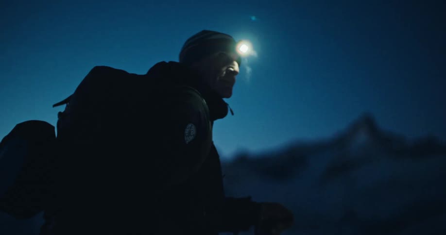 A determined mountaineer hikes through a snowy mountain landscape at night, guided by the light of his headlamp