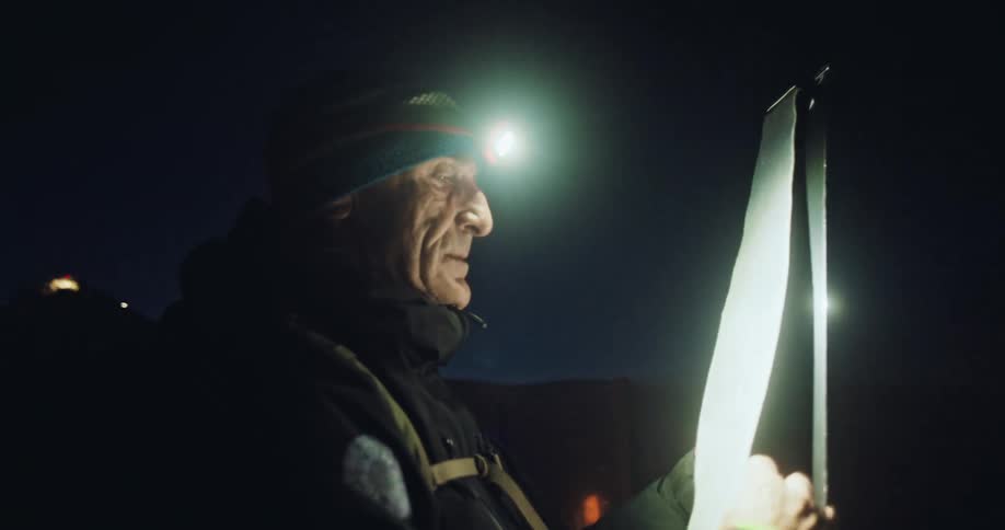 An older man wearing a headlamp carefully attaches a climbing skin to his ski in the dark before a tour