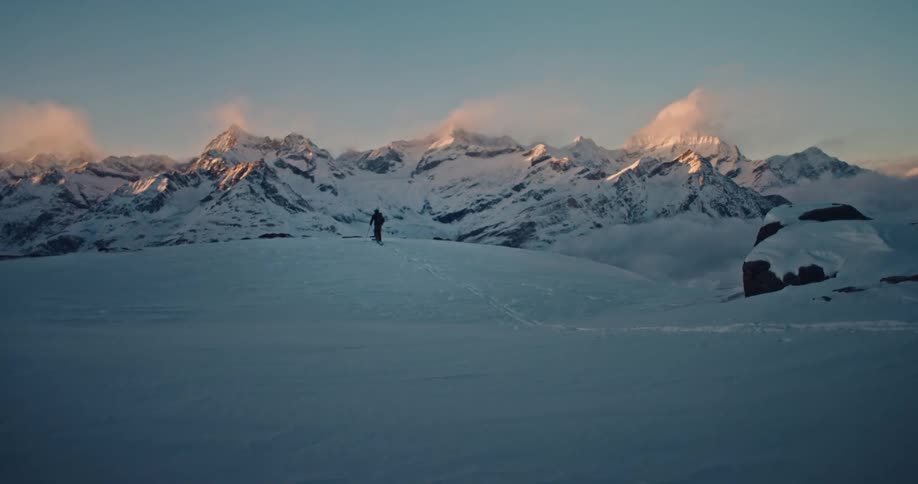 A solitary skier stands on a snowy peak, admiring the breathtaking view of a majestic mountain range at sunrise