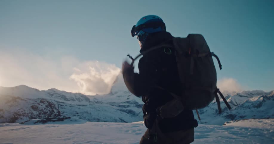 An adventurer in a helmet and goggles puts on his backpack in slow motion, preparing for an expedition with the Matterhorn in the background