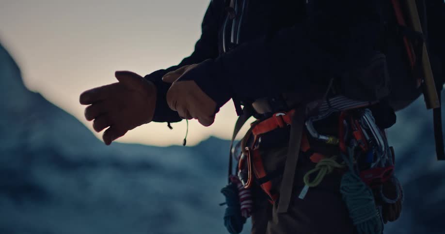 An alpinist carefully puts on their climbing gloves in slow motion with a majestic snow-capped mountain peak in the background