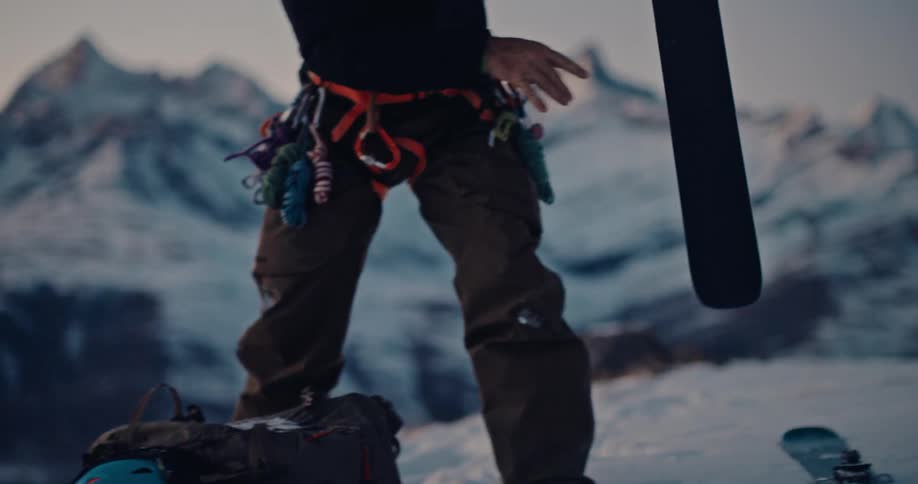 A close-up slow-motion shot shows a skier removing their skis from a backpack and placing them on the fresh snow, preparing for a descent in the mountains at sunrise