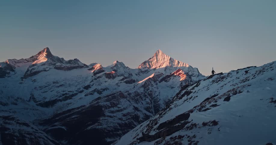 A lone skier hikes up a snowy mountain ridge towards the epic, sun-kissed alpine peaks at dawn