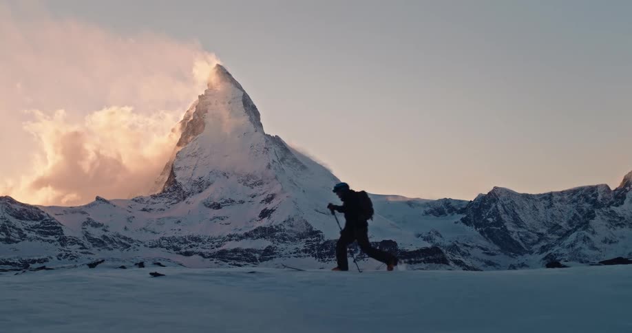 A lone ski tourer makes their way across a snowy landscape in slow motion with the iconic Matterhorn mountain in the background at sunset