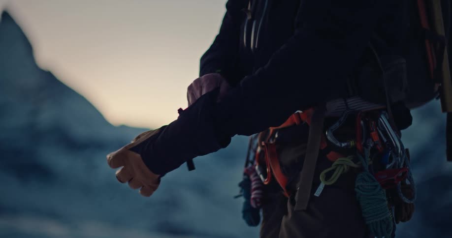 A mountain climber carefully puts on thick winter gloves in slow motion, preparing for a cold ascent with climbing gear visible
