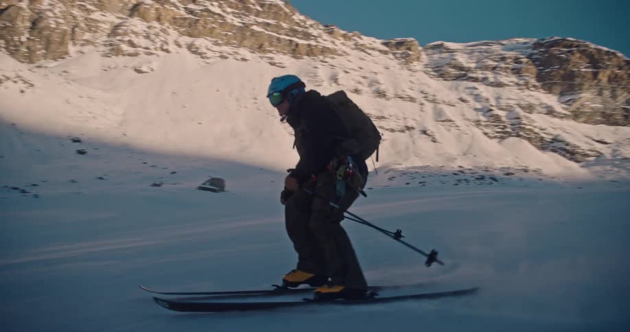 A skier carves in slow motion down a snowy mountain slope as the sun sets behind the peaks
