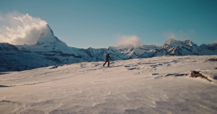 A lone skier traverses a vast, windy snowfield in the Swiss Alps, with the iconic Matterhorn mountain in the distance