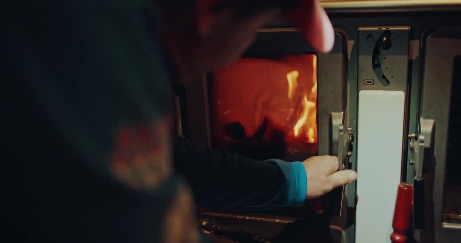 A man closes the small glass door on a traditional wood-burning stove, containing the warm, glowing fire inside