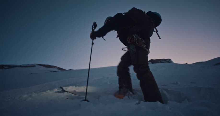 A silhouetted backcountry skier executes a kick turn on a steep, snowy slope in the blue hour, their headlamp lighting the way
