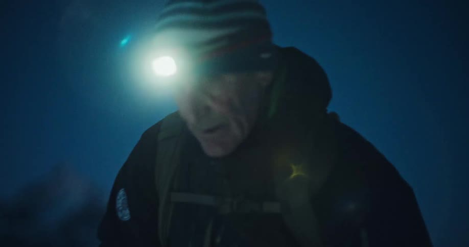 A mountaineer with a headlamp climbs a snowy mountain in the dark, with the camera slowly focusing on his determined face