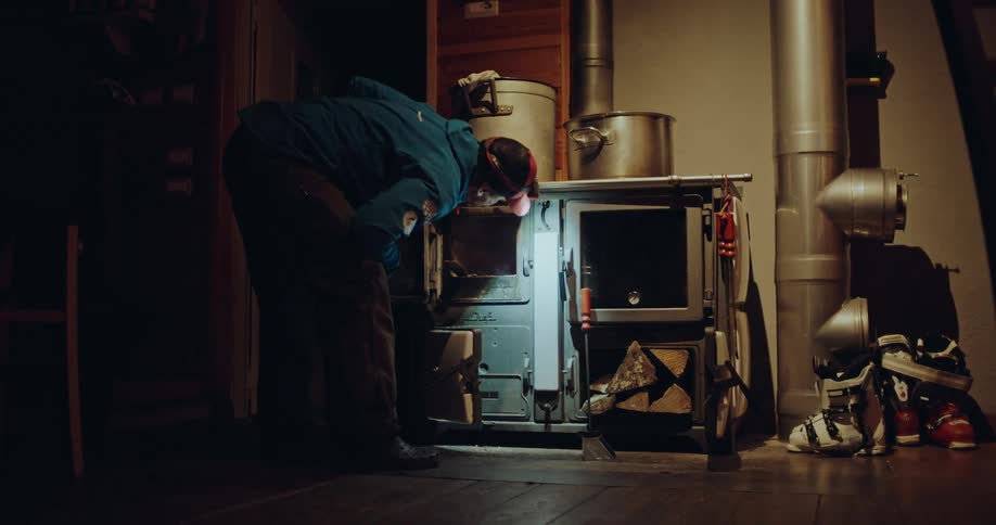 A man wearing a headlamp carefully starts a fire in a wood-burning stove inside a cozy, dark mountain hut