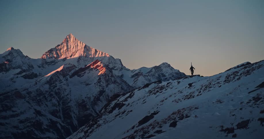 A lone backcountry skier hikes along a snowy alpine ridge at sunrise, with a majestic sunlit mountain peak in the distance