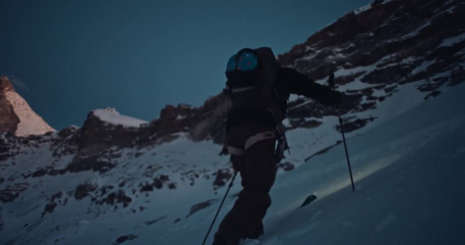 In dramatic slow motion, a mountaineer wearing a headlamp ascends a steep snowy mountain on skis during the twilight hours
