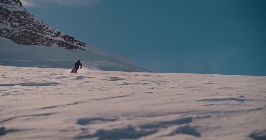 A lone skier carves through a vast, sunlit snowfield in the mountains in a beautiful slow motion shot
