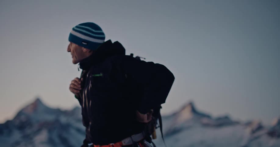 A mountaineer prepares for his expedition by packing his bag and putting it on in the snowy mountains at sunrise