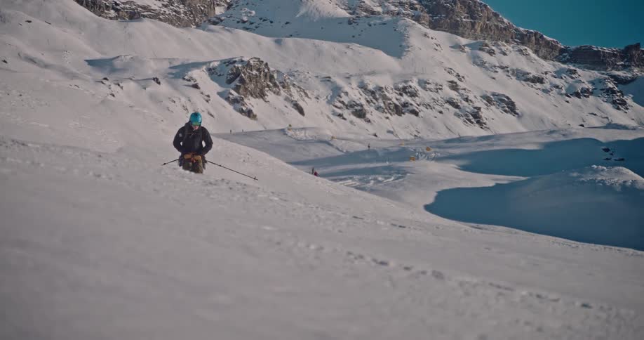Slow motion footage of a backcountry skier carving fresh tracks down a vast, sunlit mountain slope