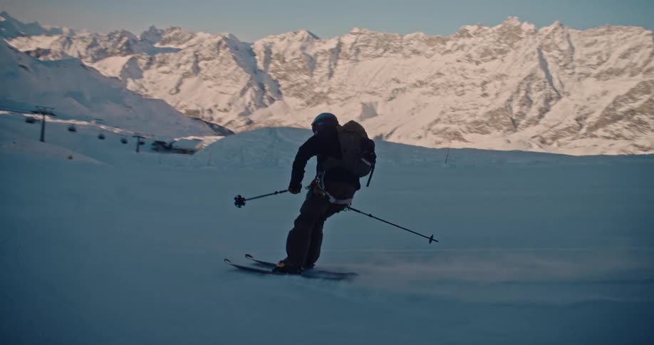 Cinematic slow motion follows a skier with a backpack gliding down a vast snowy mountain at sunset
