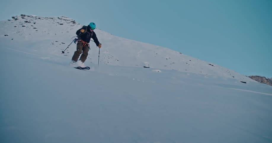 A backcountry skier makes smooth turns through fresh powder while descending a vast, sunlit mountain slope in slow motion
